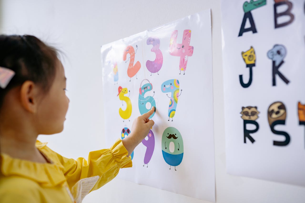 A young girl in a yellow shirt learning numbers in an educational setting, pointing at colorful numbers on a wall poster.